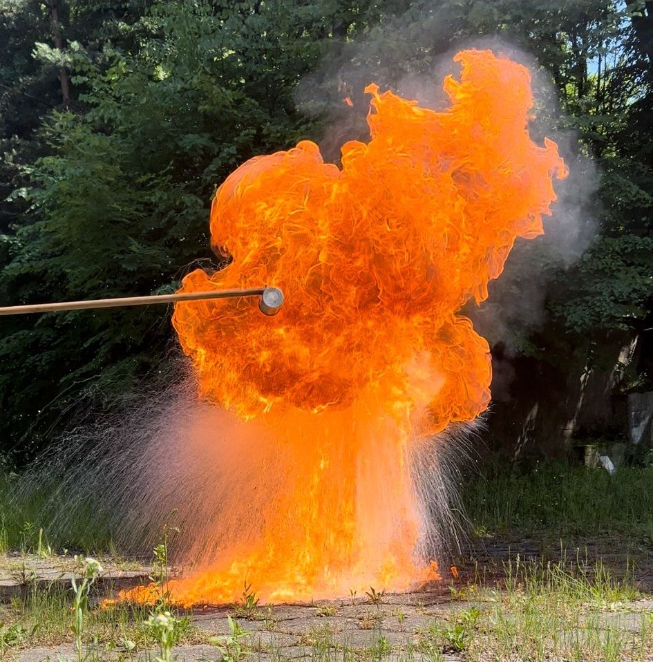 Eksplozja ognia w plenerze, wywołana prawdopodobnie kijem z metalową końcówką. Widoczne płomienie, dym i woda. Tło stanowią drzewa i fragment betonowej nawierzchni.