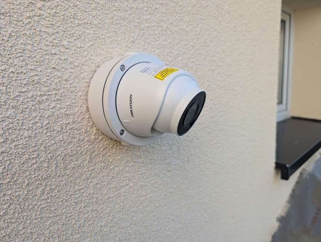 Kamera przemysłowa Hikvision, kopułkowa, zamontowana na chropowatej ścianie zewnętrznej budynku, widoczna etykieta ostrzegawcza, obok okno z czarnym parapetem.