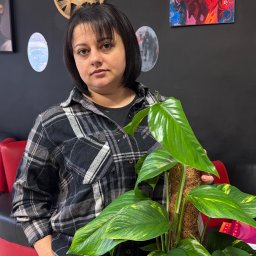 1. PIĘKNE WŁOSY U TATIANY TETIANA KOZAK 2. WARZYWNIACZEK U NAZARA TETIANA KOZAK - Kobieta z rośliną doniczkową na tle czarnej ściany z dekoracjami w Bydgoszczy. Uwagę przykuwa duża roślina z zielonymi liśćmi i brązowym palikiem.