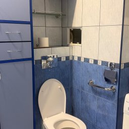 1. PIĘKNE WŁOSY U TATIANY TETIANA KOZAK 2. WARZYWNIACZEK U NAZARA TETIANA KOZAK - Niebiesko-biała łazienka po remoncie z toaletą, szafką i uchwytem dla niepełnosprawnych. Widoczne płytki i akcesoria łazienkowe. Czyste i schludne wnętrze.