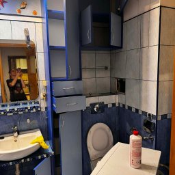 1. PIĘKNE WŁOSY U TATIANY TETIANA KOZAK 2. WARZYWNIACZEK U NAZARA TETIANA KOZAK - Niebieska łazienka z otwartymi szafkami, pralką i środkiem czyszczącym. W lustrze odbija się osoba robiąca zdjęcie. Widoczne muszelki jako dekoracja.