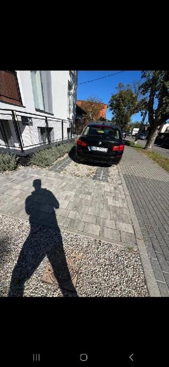 Ułożona kostka brukowa na podjeździe do domu, z zaparkowanym czarnym samochodem. Widoczny cień osoby. Słoneczny dzień, błękitne niebo.