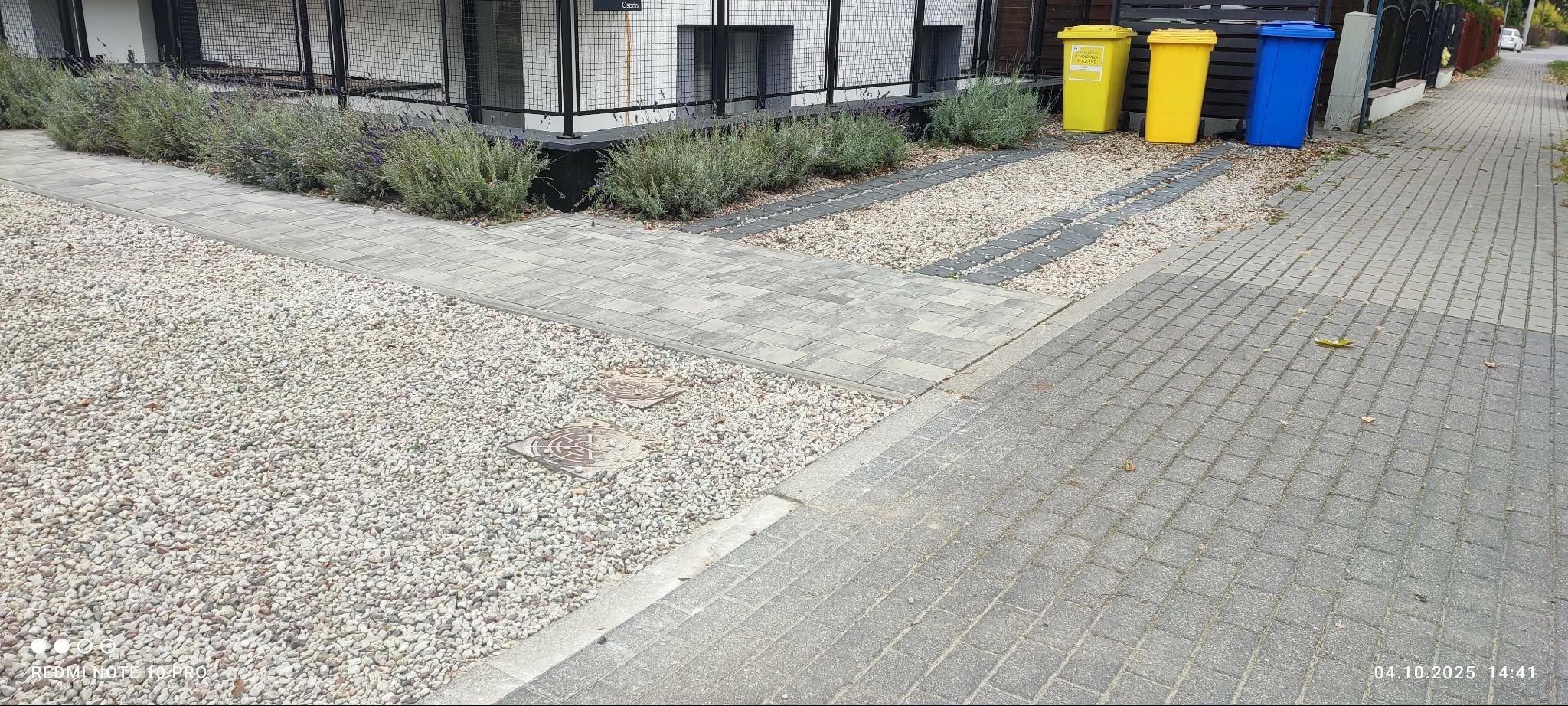 Ułożona kostka brukowa i kamień ozdobny tworzą ścieżkę w ogrodzie. Widoczne rośliny, budynek z czarną balustradą i kolorowe pojemniki na śmieci.