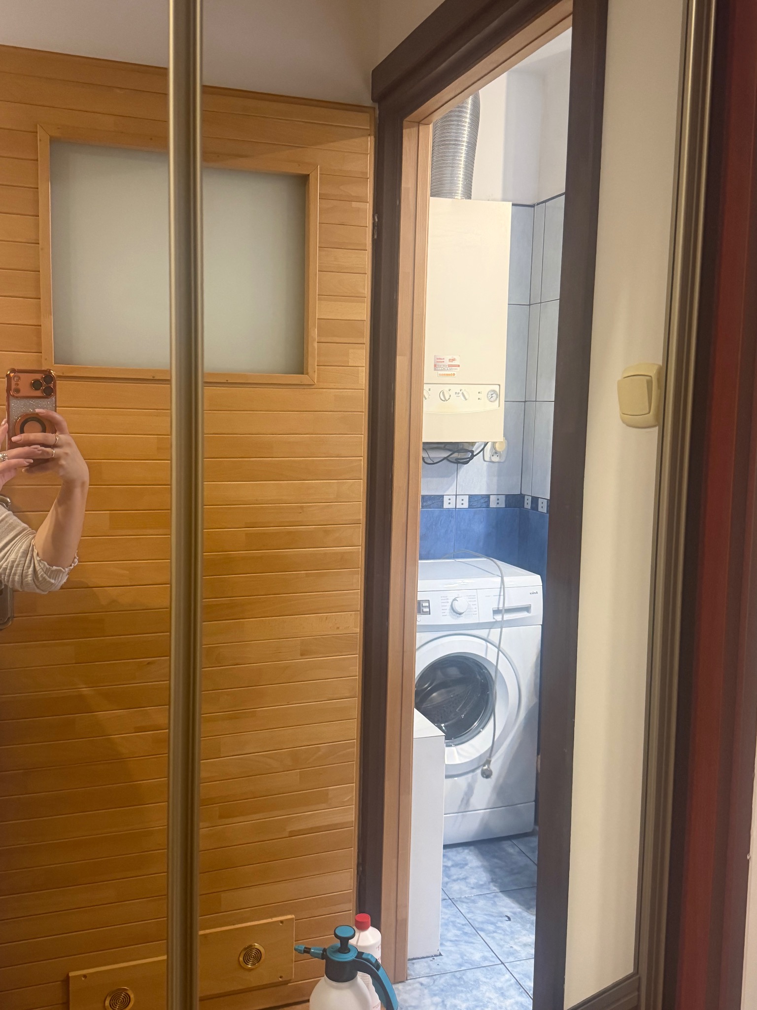 Widok na łazienkę z pralką i piecem gazowym przez otwarte drzwi szafy wnękowej w Bydgoszczy. W odbiciu widać osobę robiącą zdjęcie.