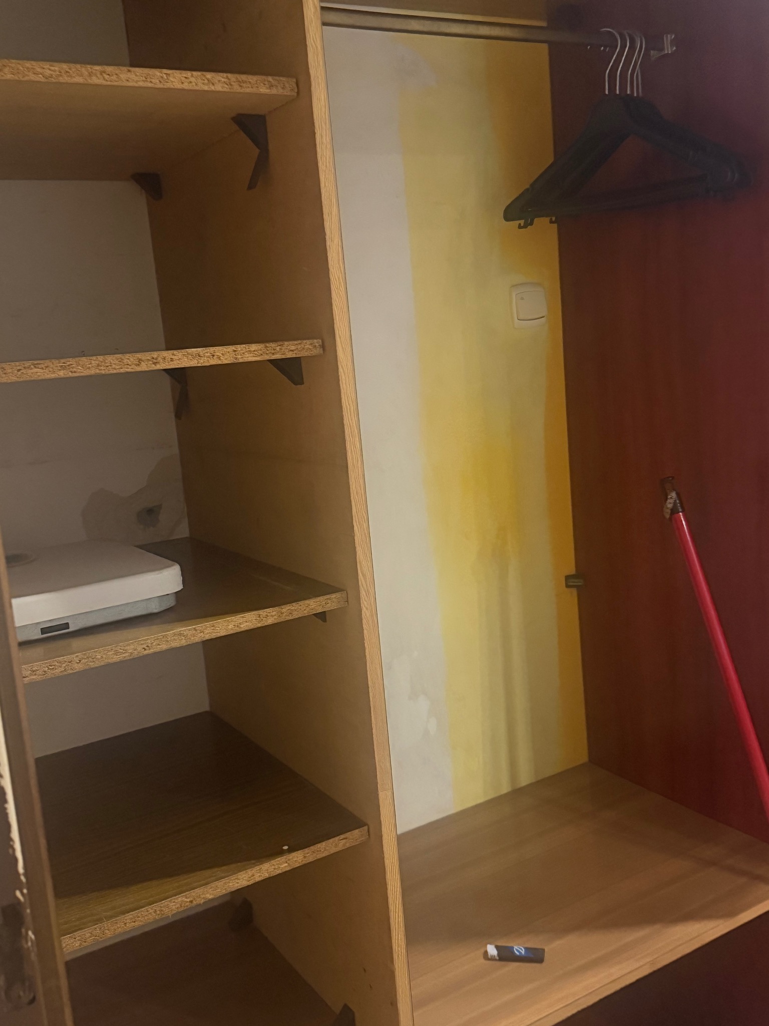 Wnętrze szafy z półkami i wieszakami, widoczne ślady po zalaniu na ścianie, na dolnej półce leży mały przedmiot. Szafa w odcieniach brązu i czerwieni.