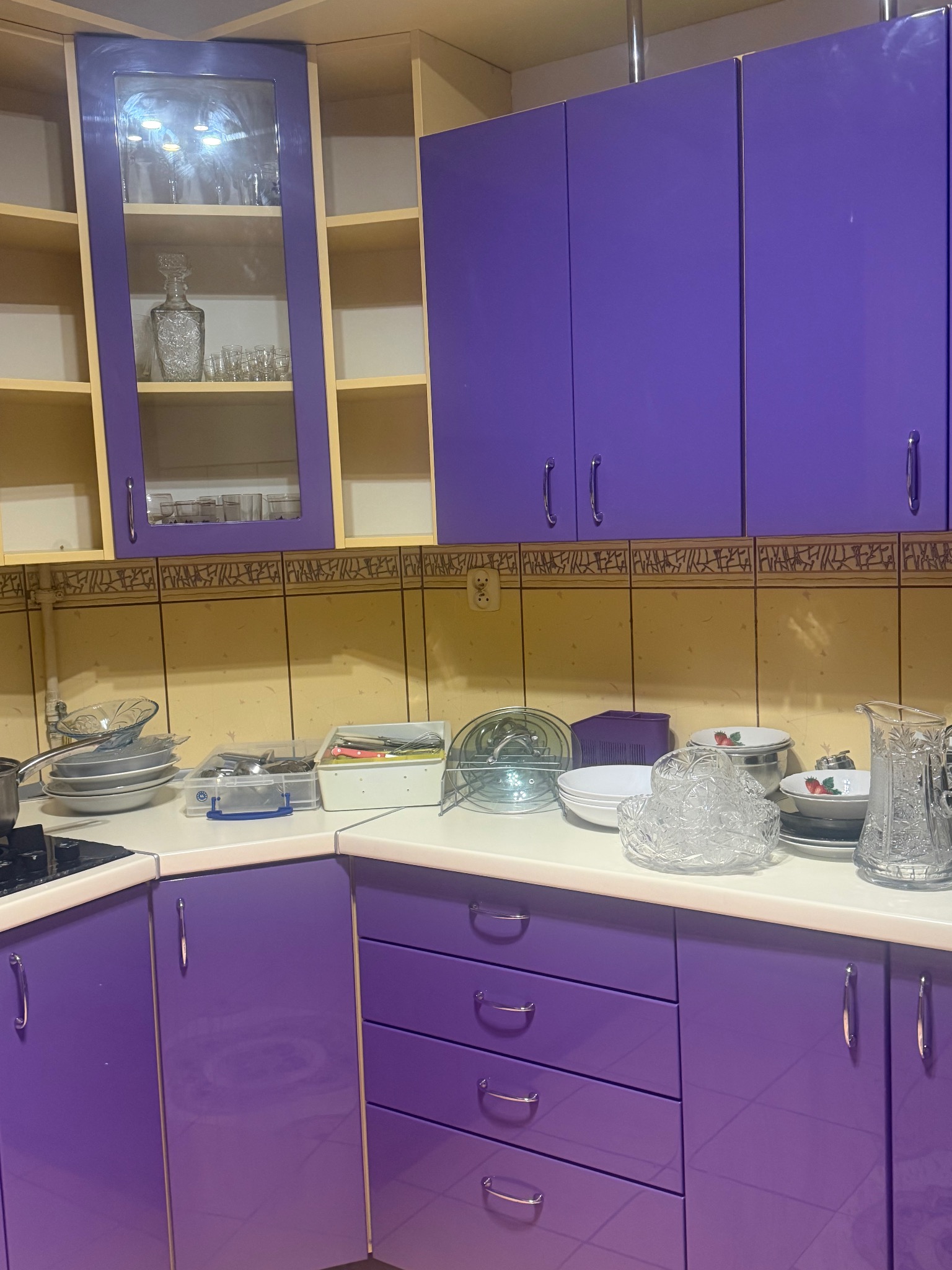 Fioletowa kuchnia z naczyniami na blacie. Widoczne szafki górne z przeszklonymi drzwiczkami i dolne z szufladami. Na blacie naczynia, sztućce i akcesoria kuchenne.