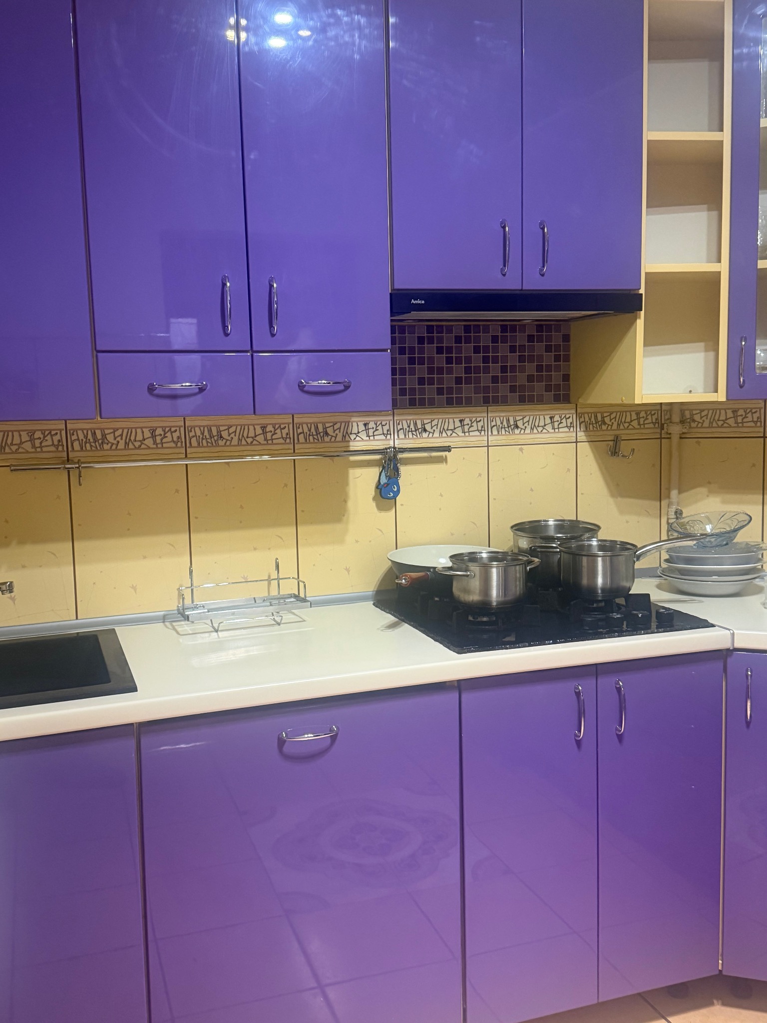 Fioletowa zabudowa kuchenna z błyszczącymi frontami, na blacie płyta grzewcza z garnkami i naczyniami. Nad blatem dekoracyjne płytki i szafki wiszące.