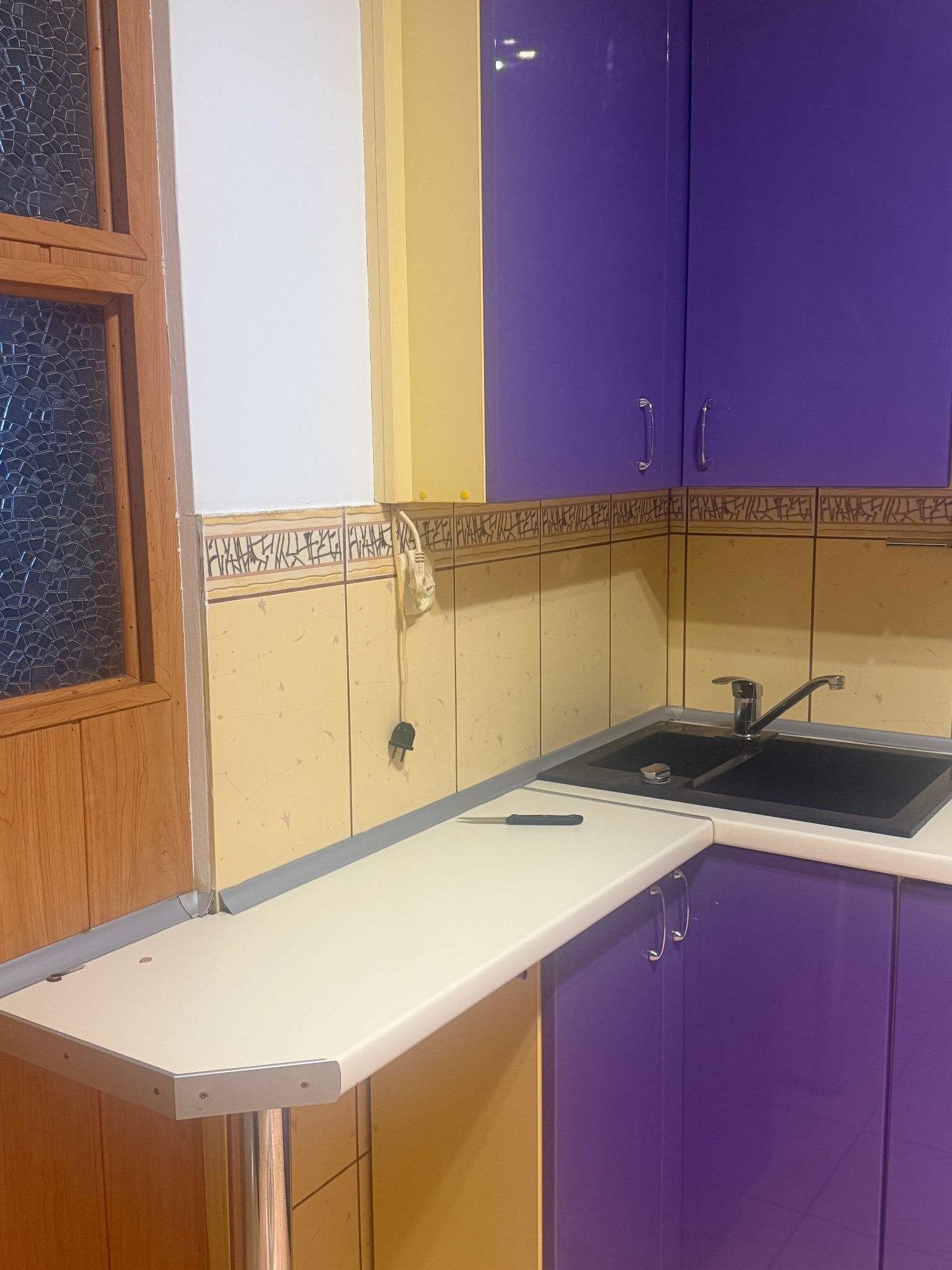 Kuchnia z fioletowymi szafkami i żółtymi płytkami. Na blacie leży nóż. Widoczne gniazdko elektryczne i okno z ozdobną szybą.