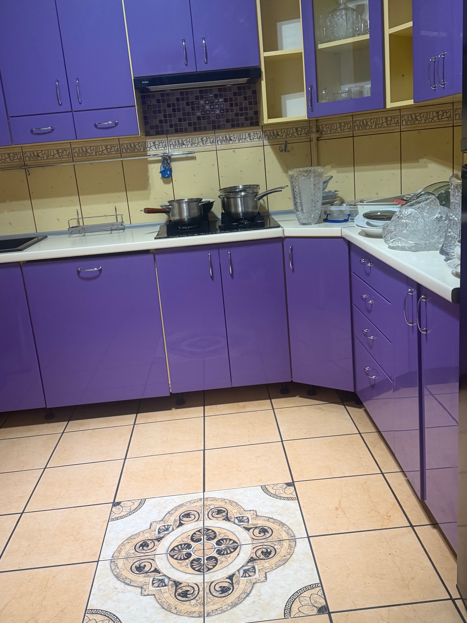 Fioletowa kuchnia z beżowymi płytkami i wzorzystą podłogą. Na blacie garnki na płycie, naczynia i kryształowy wazon. Ujęcie z niskiego kąta.