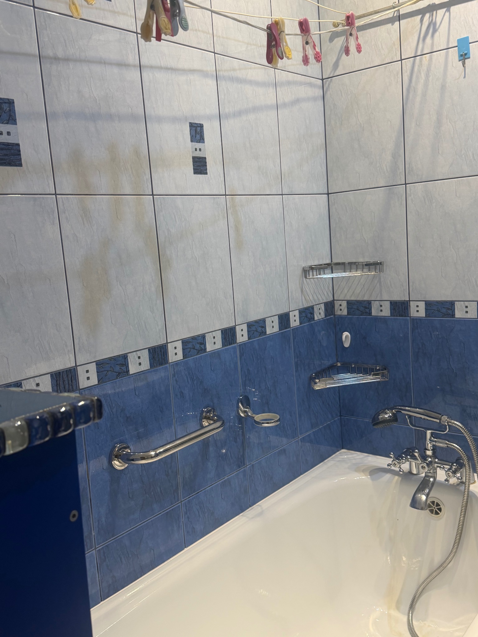 Biała wanna z chromowaną armaturą na tle niebiesko-białych płytek. Na ścianie uchwyt, półki i suszarka z klamerkami. Widoczne ślady użytkowania na fugach.