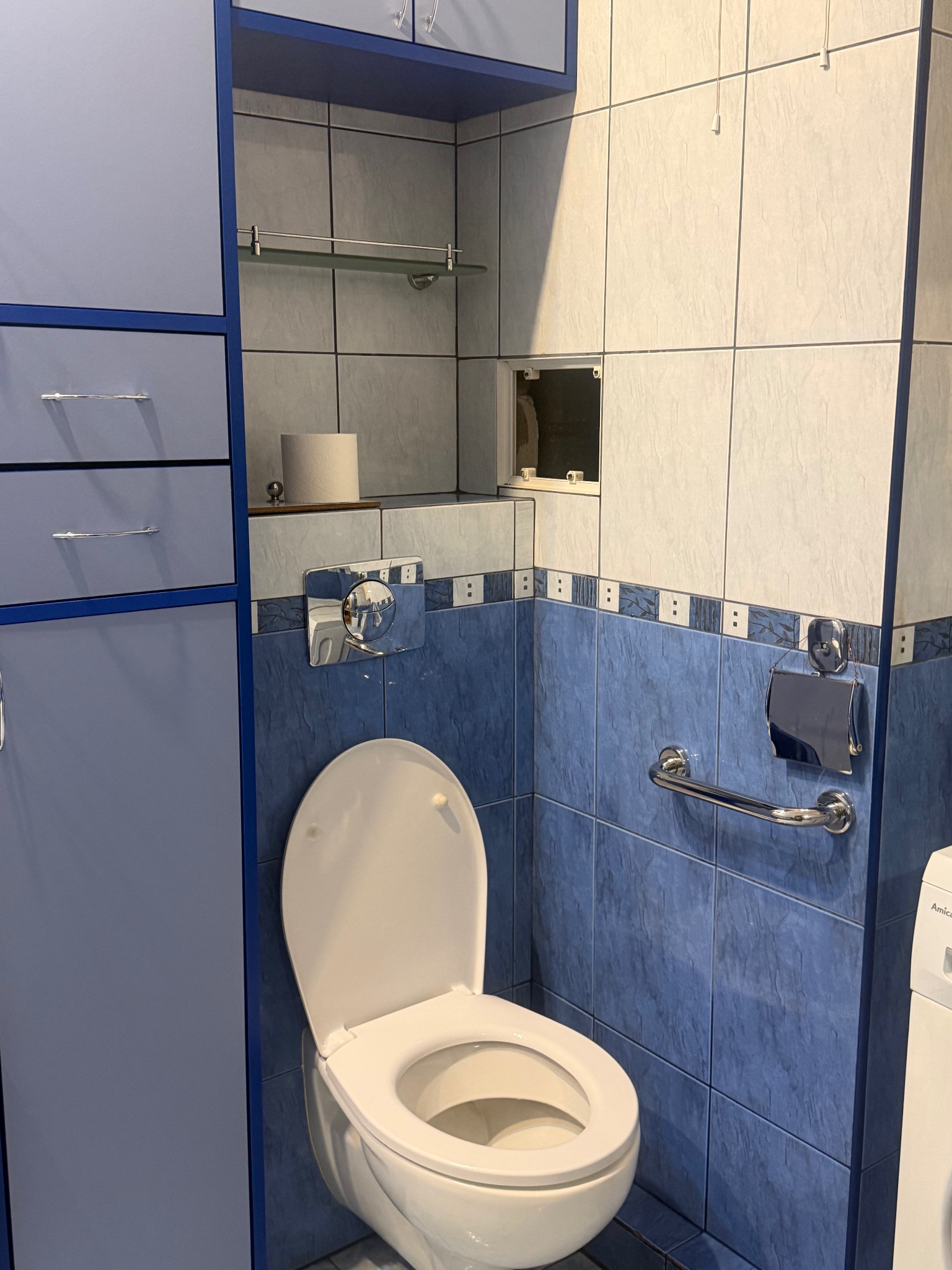 Niebiesko-biała łazienka po remoncie z toaletą, szafką i uchwytem dla niepełnosprawnych. Widoczne płytki i akcesoria łazienkowe. Czyste i schludne wnętrze.