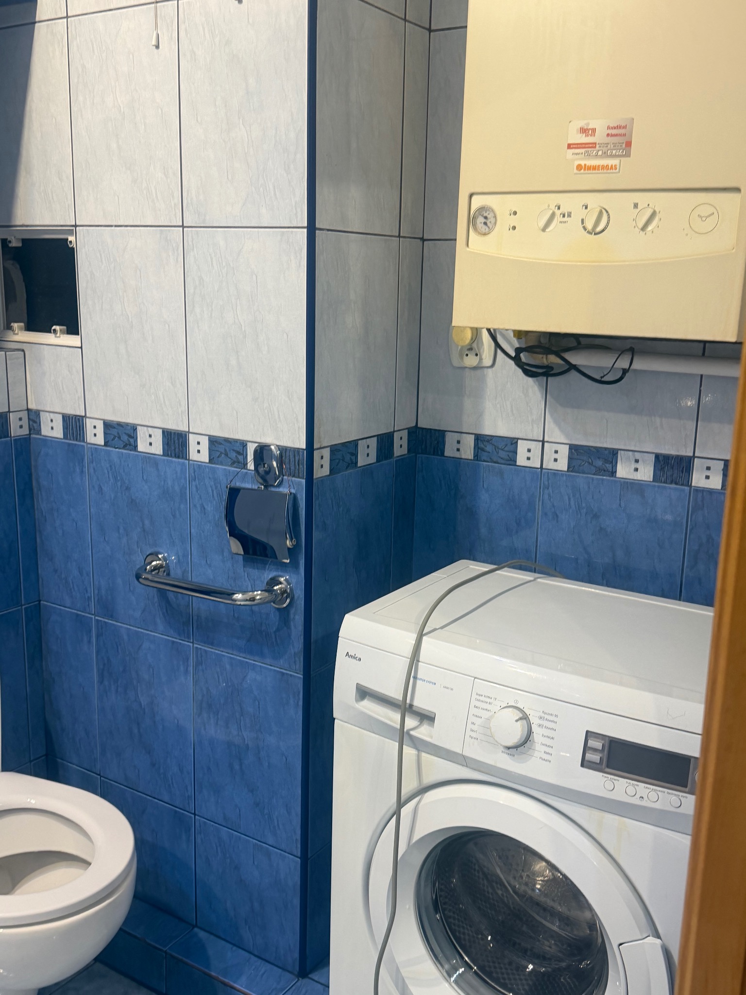 Łazienka z niebieskimi płytkami, białą pralką Amica i piecem gazowym Immergas. Widoczny fragment toalety i uchwyt na papier. Ujęcie z bliska, bez wyraźnego oświetlenia.