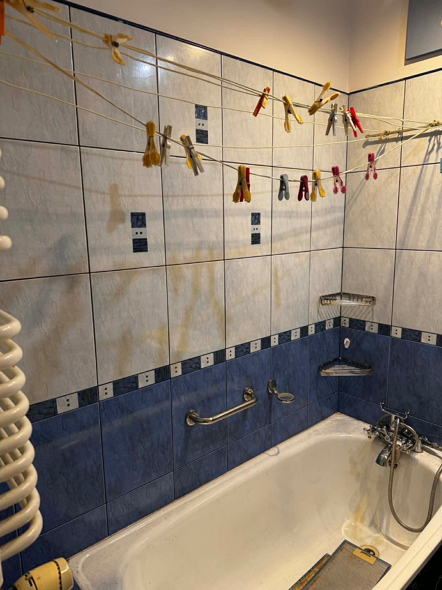 Łazienka z wanną, na której suszą się rzeczy na sznurku z klamerkami. Widoczne płytki na ścianach i armatura łazienkowa.