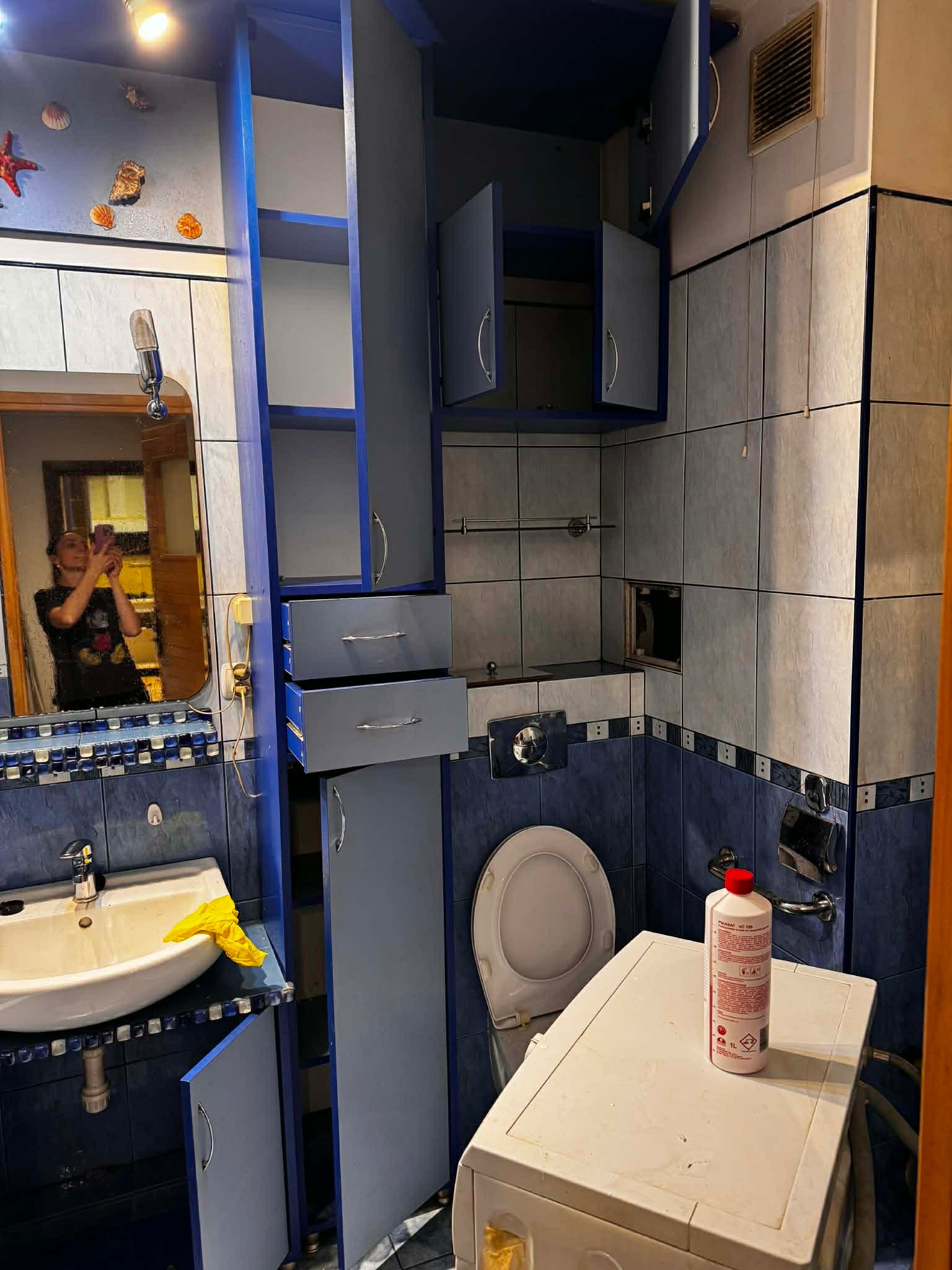 Niebieska łazienka z otwartymi szafkami, pralką i środkiem czyszczącym. W lustrze odbija się osoba robiąca zdjęcie. Widoczne muszelki jako dekoracja.