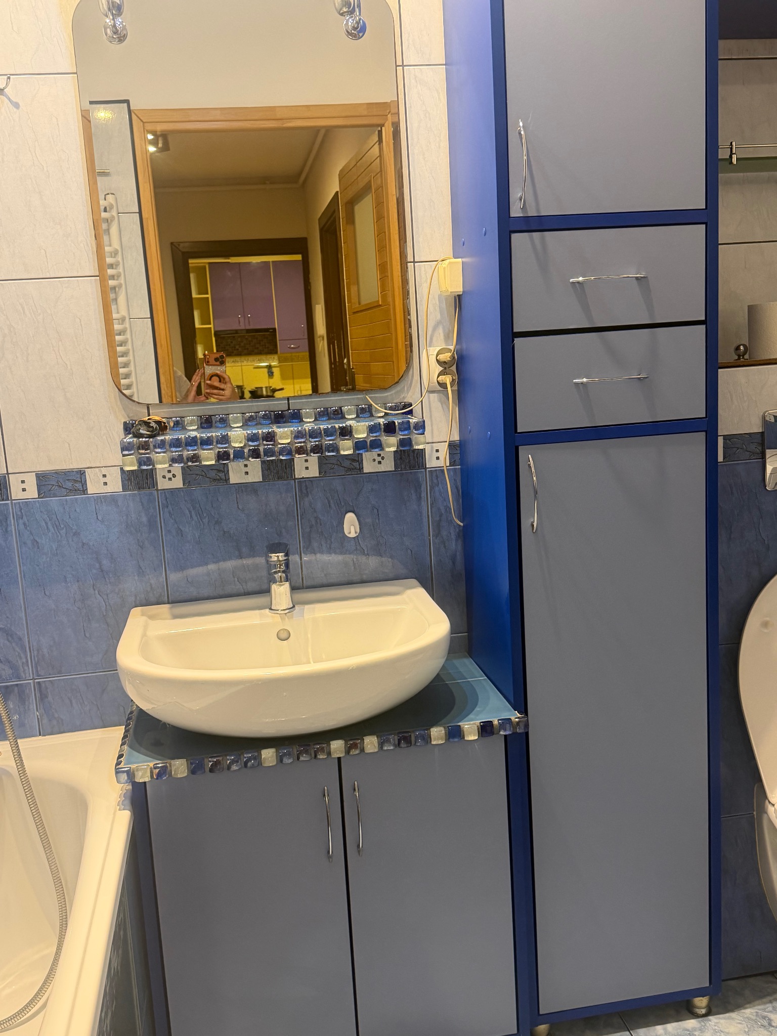 Niebiesko-szara łazienka z umywalką i szafkami, lustro odbija fragment kuchni. Dekoracyjne mozaiki dodają unikalnego charakteru wnętrzu.