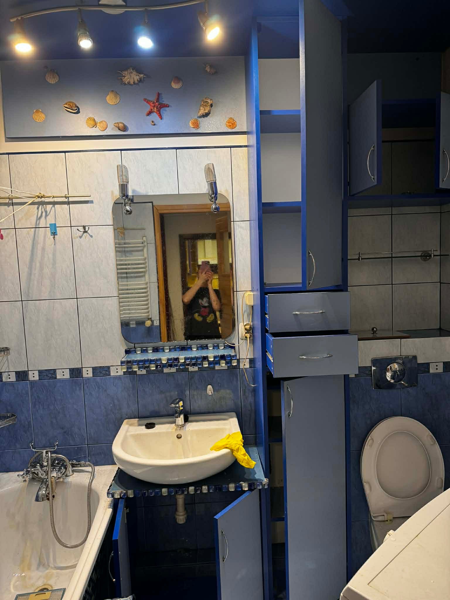 Niebieska łazienka z umywalką, toaletą i szafkami. W lustrze odbija się osoba robiąca zdjęcie. Dekoracje z muszelek na ścianie. Widoczna żółta rękawica.