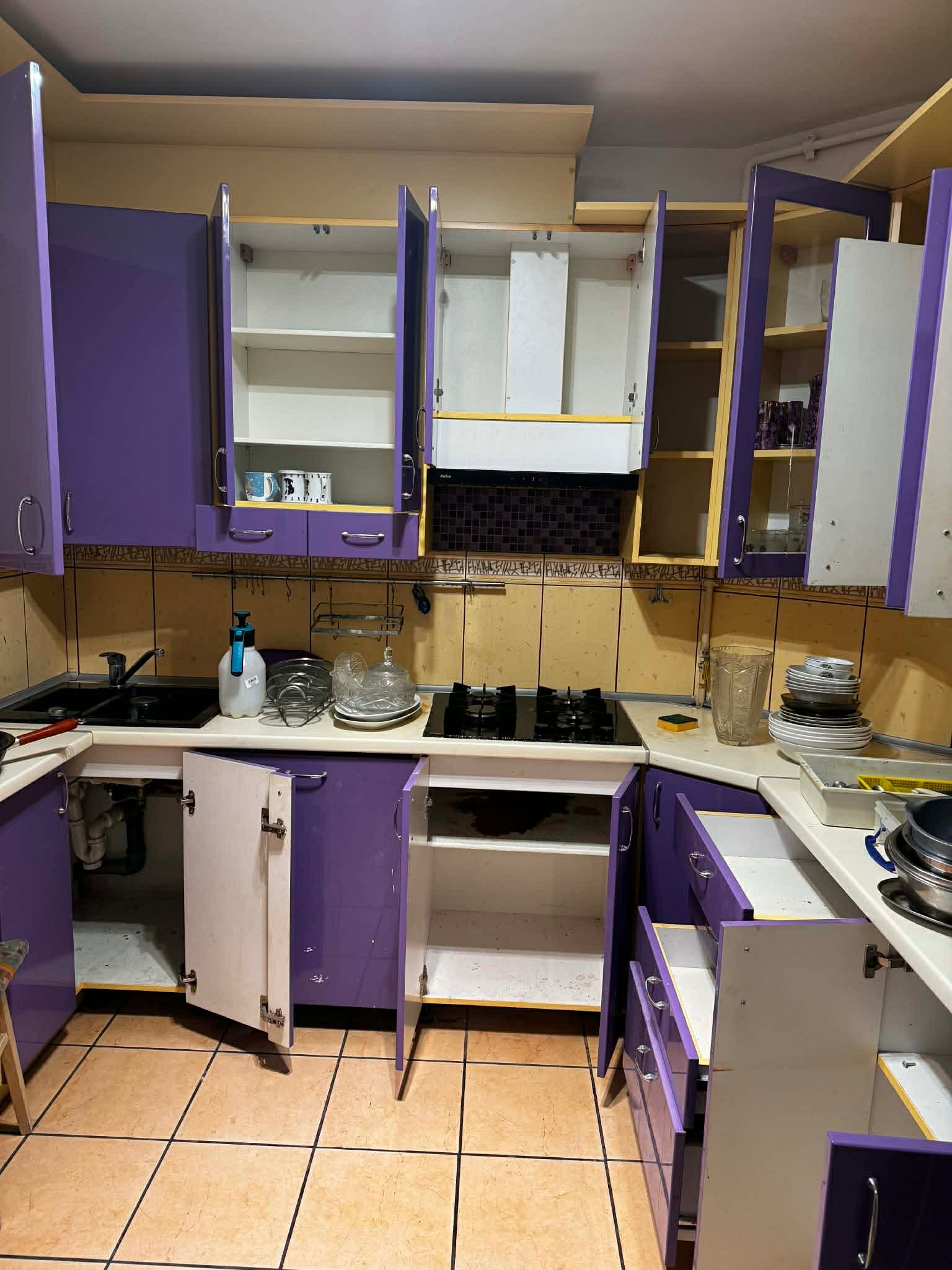 Niefotogeniczna kuchnia z otwartymi szafkami i szufladami, naczyniami na blacie i butelką płynu do mycia. Widoczne płytki na ścianie i podłodze.