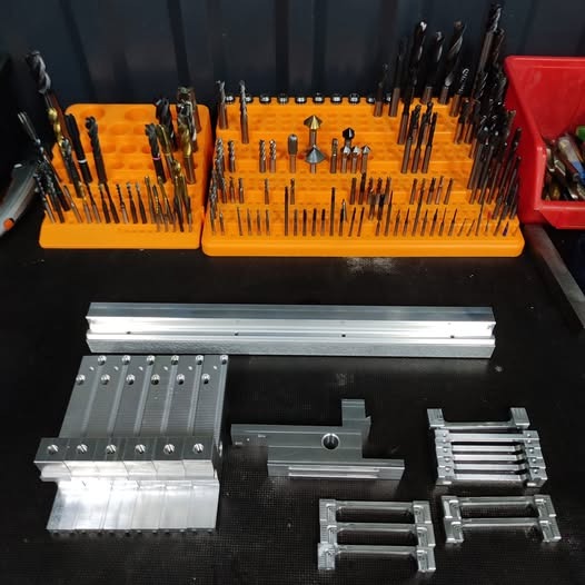 Precyzyjnie obrobione metalowe elementy na blacie warsztatowym, obok wiertła w pomarańczowych organizerach. Detaliczna prezentacja efektów obróbki.