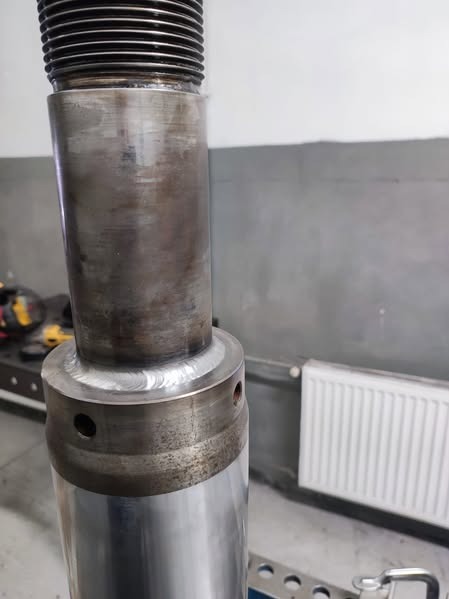 Precyzyjnie wykonany spaw na metalowym cylindrze z gwintowaną górą, widoczne ślady obróbki i szlifowania, surowe wnętrze warsztatu w tle.