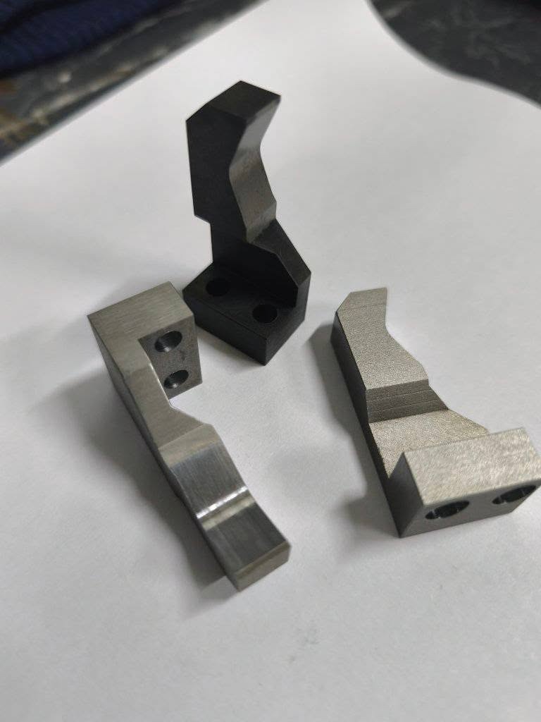 Precyzyjnie wykonane, metalowe elementy o nieregularnych kształtach, widoczne ślady obróbki. Detale z otworami, ułożone na jasnym tle. Różne wykończenia powierzchni.