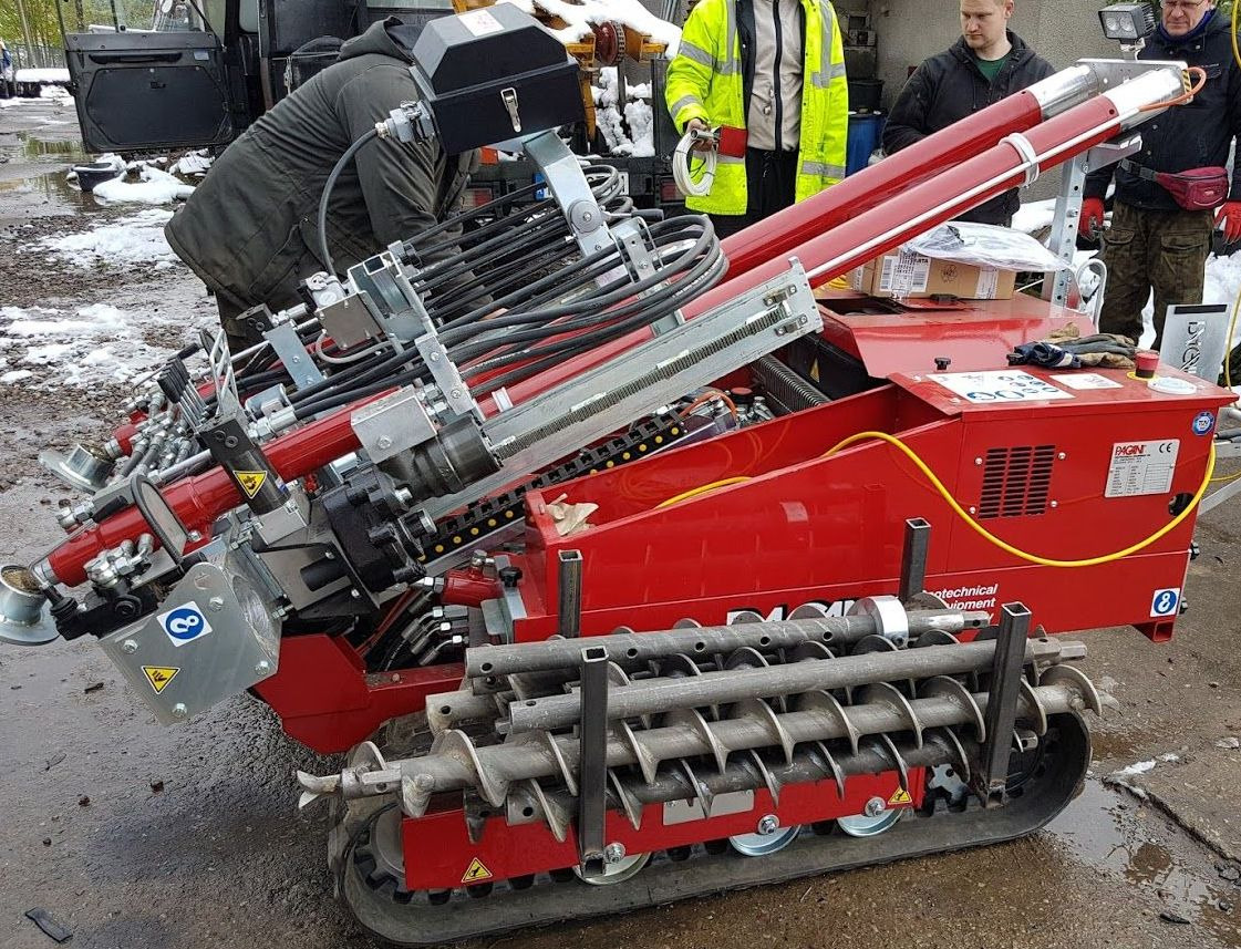 Czerwona wiertnica geologiczna na gąsienicach w trakcie pracy, widoczni operatorzy, śnieg na ziemi. Sprzęt do odwiertów geologicznych.