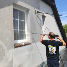 Pracownik w koszulce z logo firmy czyści elewację budynku myjką ciśnieniową, widoczne okno z ceglanym parapetem i fragment dachu. Częściowo umyta ściana.
