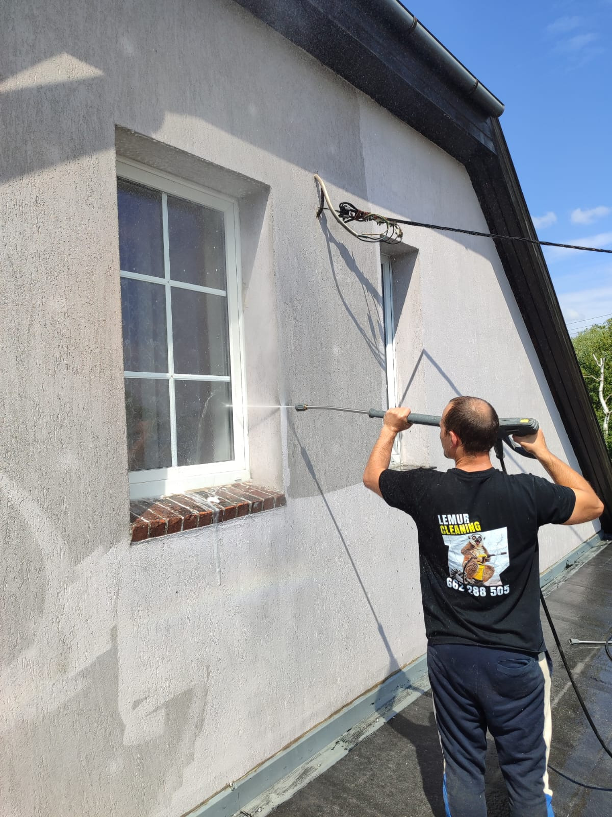 Pracownik w koszulce z logo firmy czyści elewację budynku myjką ciśnieniową, widoczne okno z ceglanym parapetem i fragment dachu. Częściowo umyta ściana.