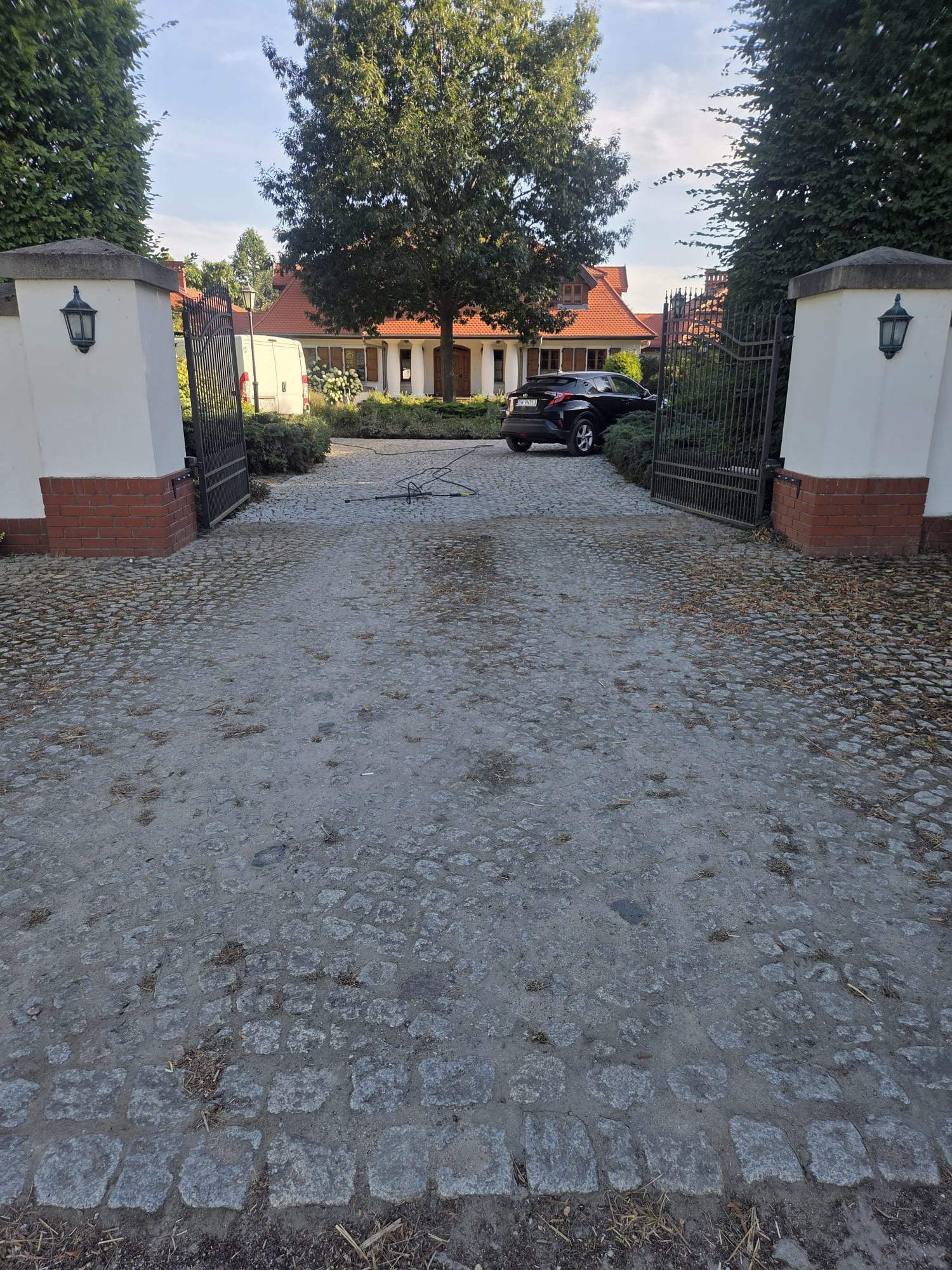 Zabrudzona kostka brukowa na podjeździe do domu z otwartą bramą, samochodem i wężem ciśnieniowym. Widoczne ślady użytkowania i zaniedbania przed czyszczeniem.