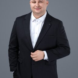 Szymon Ponikwia - Portret uśmiechniętego mężczyzny w ciemnym garniturze i białej koszuli na szarym tle. Profesjonalny wizerunek, budzący zaufanie, idealny dla doradcy.