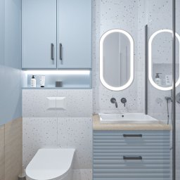 Noya - Niebieska szafka nad toaletą i umywalką z szufladami w łazience wykończonej w stylu lastryko. Owalne lustra z podświetleniem LED i drewniana podłoga.