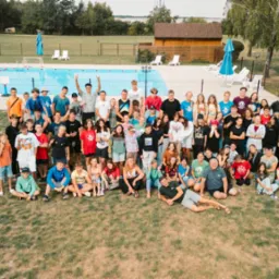 Grupowe zdjęcie uczestników kolonii lub obozu na trawie, z basenem i budynkami w tle, wykonane w słoneczny dzień. Wszyscy uśmiechnięci i zadowoleni.