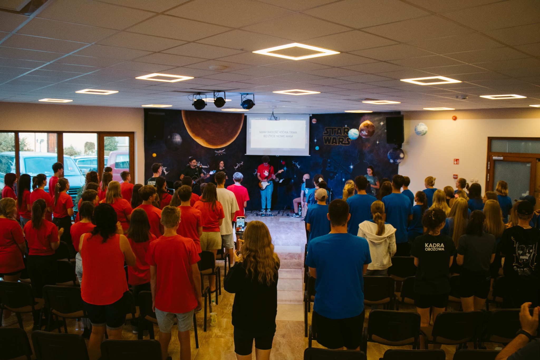 Wieczorne spotkanie na obozie: grupa młodzieży w czerwonych i niebieskich koszulkach śpiewa z zespołem na tle sceny z motywami kosmicznymi i napisem Star Wars.