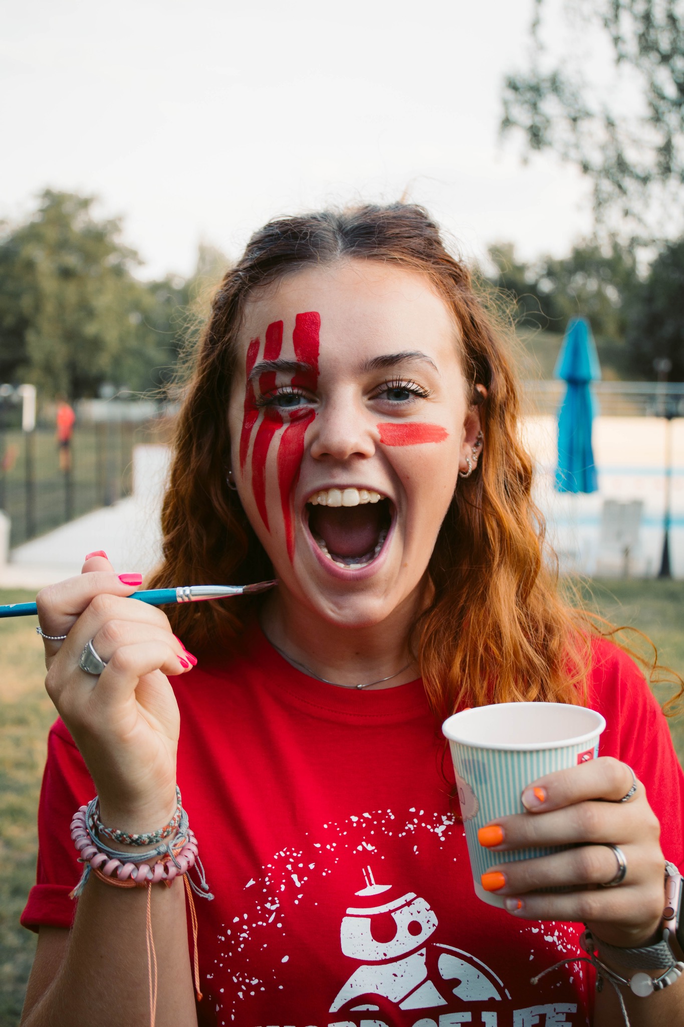 Uśmiechnięta, ruda dziewczyna z pomalowaną twarzą trzyma pędzel i kubek. W tle basen i drzewa. Atrakcja kolonijna: malowanie twarzy.