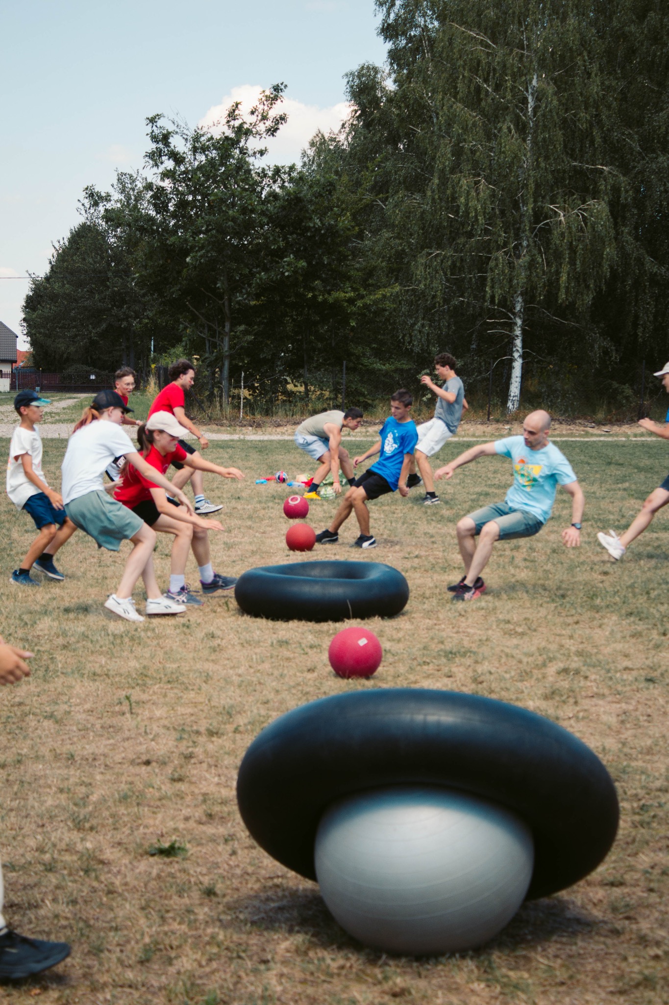 Młodzież gra w grę zespołową na trawie z użyciem opon i piłek. Uczestnicy w ruchu, w tle drzewa i budynek. Aktywność na świeżym powietrzu podczas obozu.