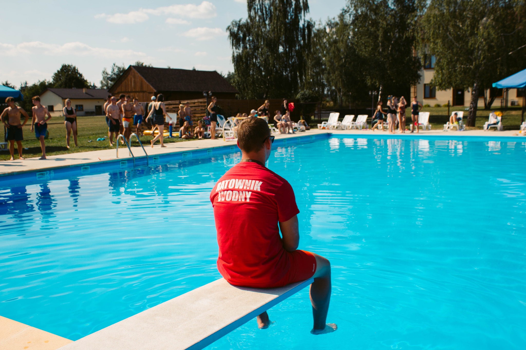 Ratownik Wodny w czerwonej koszulce siedzi na trampolinie basenu w Zgierzu, obserwując grupę osób w tle. Scena z kolonii lub obozu.