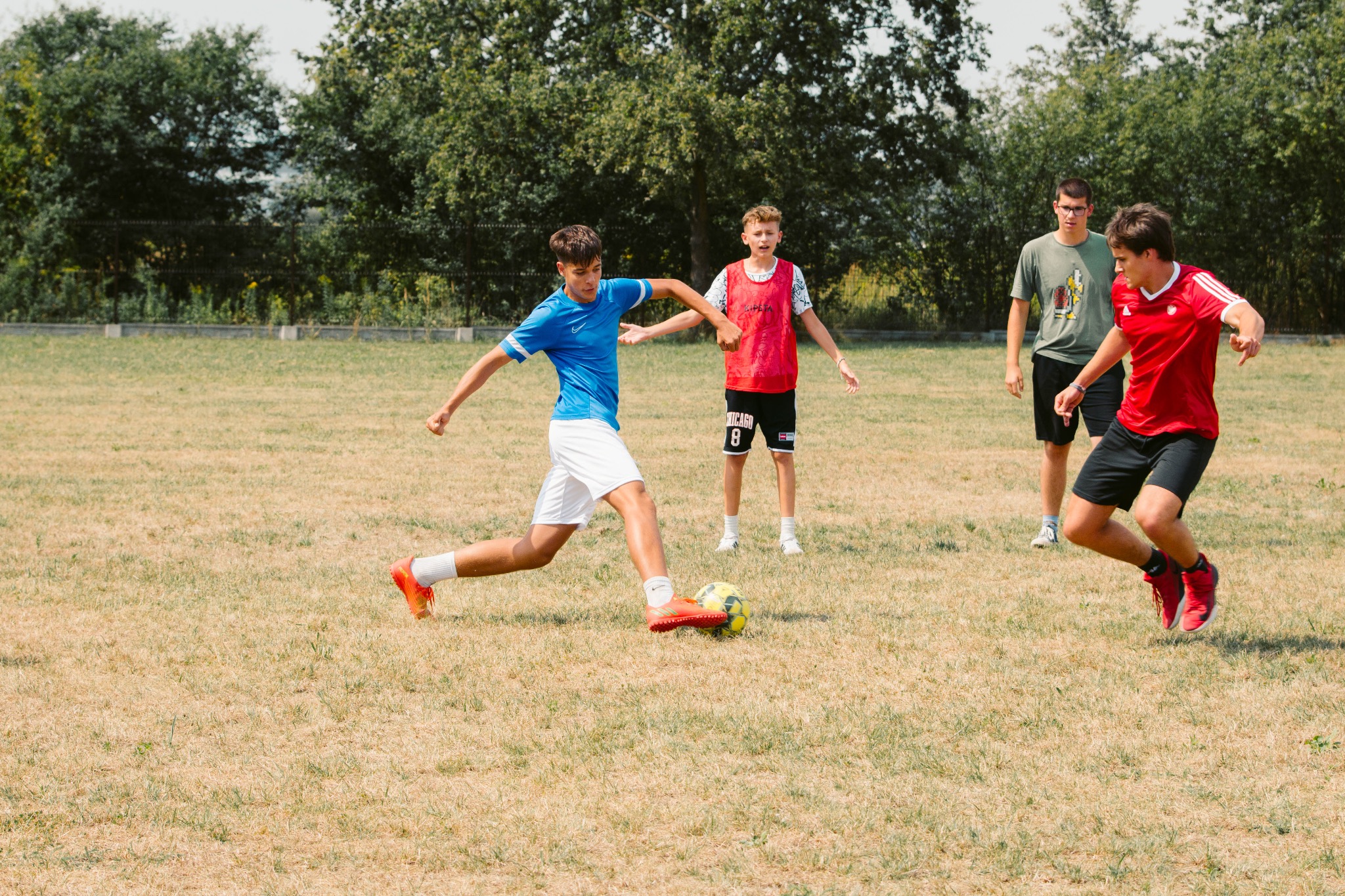 Chłopiec w niebieskiej koszulce kopie piłkę podczas treningu na trawiastym boisku. W tle drzewa i obserwujący koledzy.