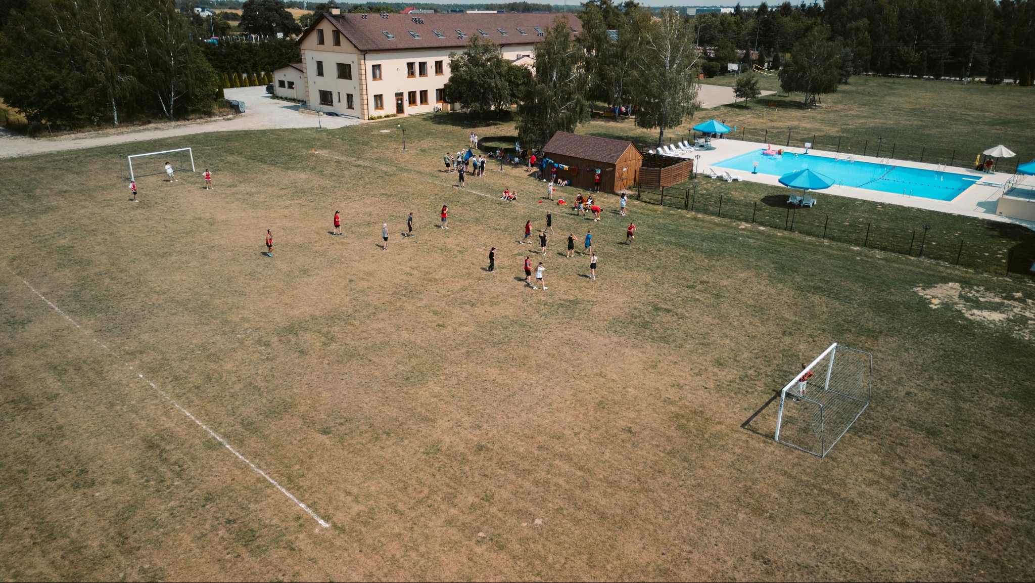 Widok z lotu ptaka na ośrodek kolonijny: boisko z młodzieżą grającą w piłkę nożną, budynek, basen z parasolami i leżakami. Letni dzień.