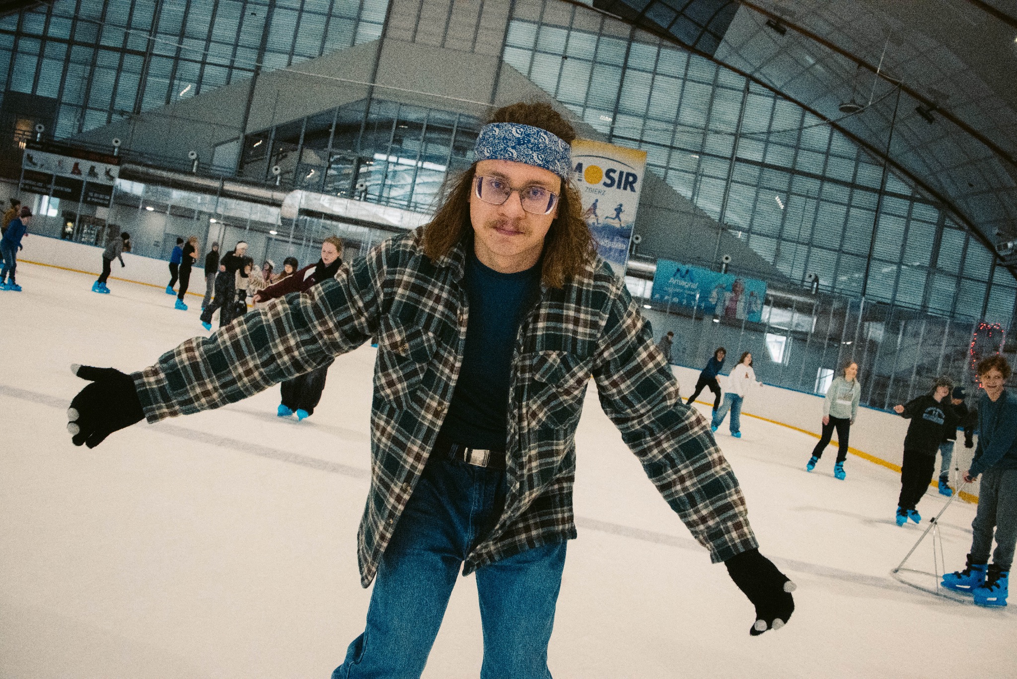 Mężczyzna na lodowisku w koszuli w kratę i opasce, z rozłożonymi rękami. W tle inni uczestnicy obozu na łyżwach i banery reklamowe.