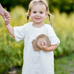 Klaudia Pociask Fotografia - Uśmiechnięta dziewczynka w białej sukience trzyma drewniany aparat, stojąc na tle zieleni, prowadzona za rękę przez osobę z manicure.