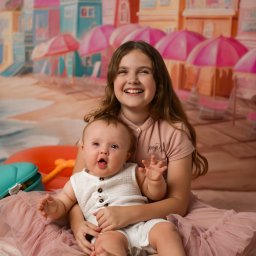 Klaudia Pociask Fotografia - Uśmiechnięta nastolatka obejmuje siedzącego na jej kolanach roześmianego niemowlaka na tle kolorowej plaży z parasolami i budynkami. Sesja w studio.