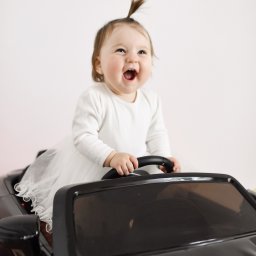 Klaudia Pociask Fotografia - Uśmiechnięta dziewczynka w białej sukience siedzi za kierownicą czarnego, zabawkowego samochodu na jasnym tle. Dziecko ma włosy związane w mały kucyk.