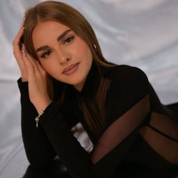 Klaudia Pociask Fotografia - Portret kobiety w czarnym body z siateczkowymi wstawkami, leżącej na błyszczącej powierzchni z rękami pod głową, na tle białej, drapowanej tkaniny.