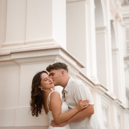 Klaudia Pociask Fotografia - Zakochana para przytulona na tle klasycznej architektury z łukami. Mężczyzna całuje kobietę w policzek. Ona w białej sukience, on w jasnej koszuli.