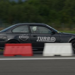 Klaudia Pociask Fotografia - Czarny samochód sportowy BMW E36 z naklejkami sponsorskimi, mknący po torze wyścigowym obok czerwono-białych barier. Kierowca w kasku widoczny wewnątrz.