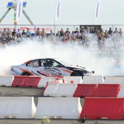 Klaudia Pociask Fotografia - Driftingowy samochód w akcji, otoczony dymem na tle trybun pełnych widzów i banerów reklamowych. Dynamiczne ujęcie z bliska, z widocznymi detalami bolidu.