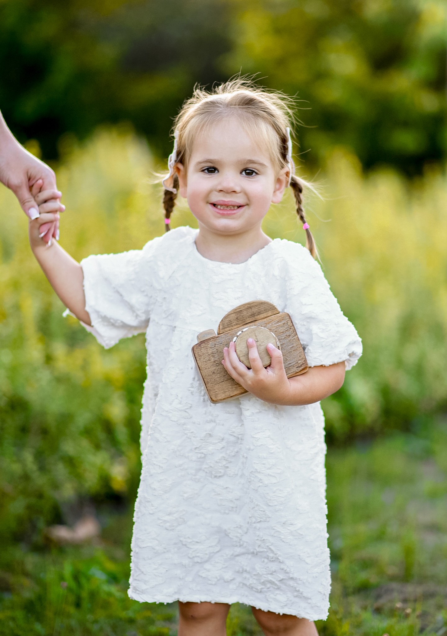 Uśmiechnięta dziewczynka w białej sukience trzyma drewniany aparat, stojąc na tle zieleni, prowadzona za rękę przez osobę z manicure.