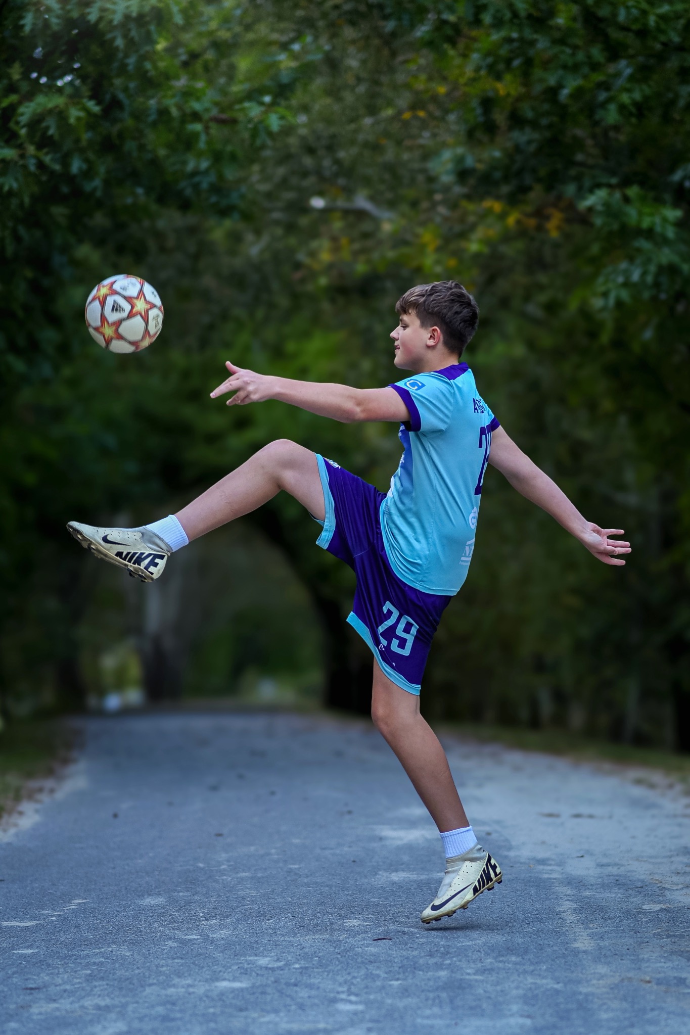 Chłopiec w sportowym stroju, kopie piłkę na drodze otoczonej drzewami. Widoczne buty Nike i numer 29 na spodenkach. Dynamiczne ujęcie w plenerze.