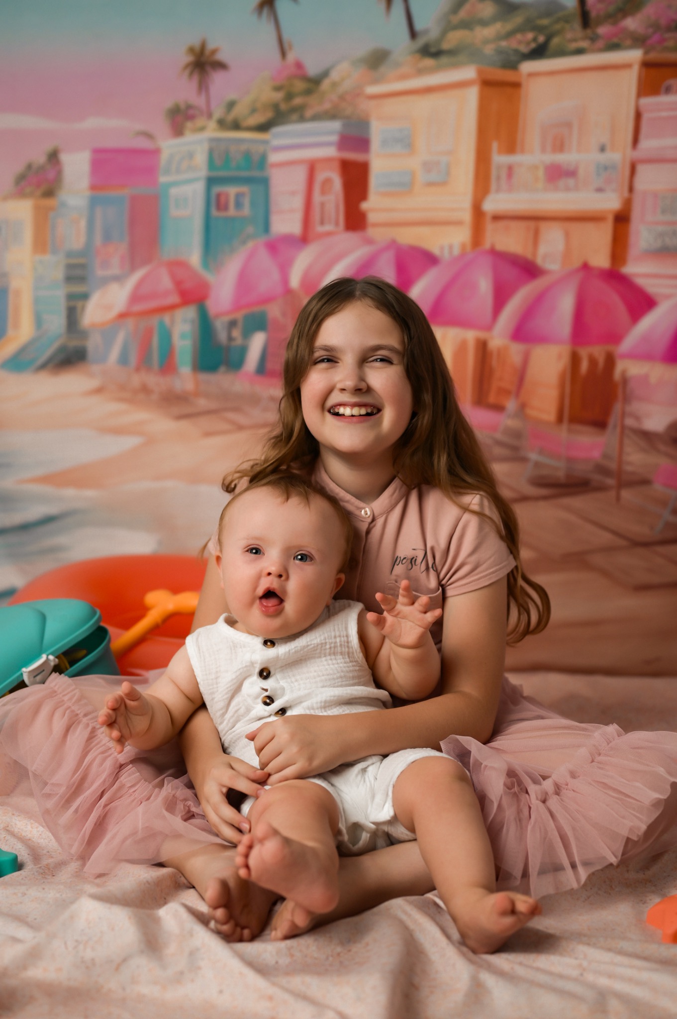 Uśmiechnięta nastolatka obejmuje siedzącego na jej kolanach roześmianego niemowlaka na tle kolorowej plaży z parasolami i budynkami. Sesja w studio.