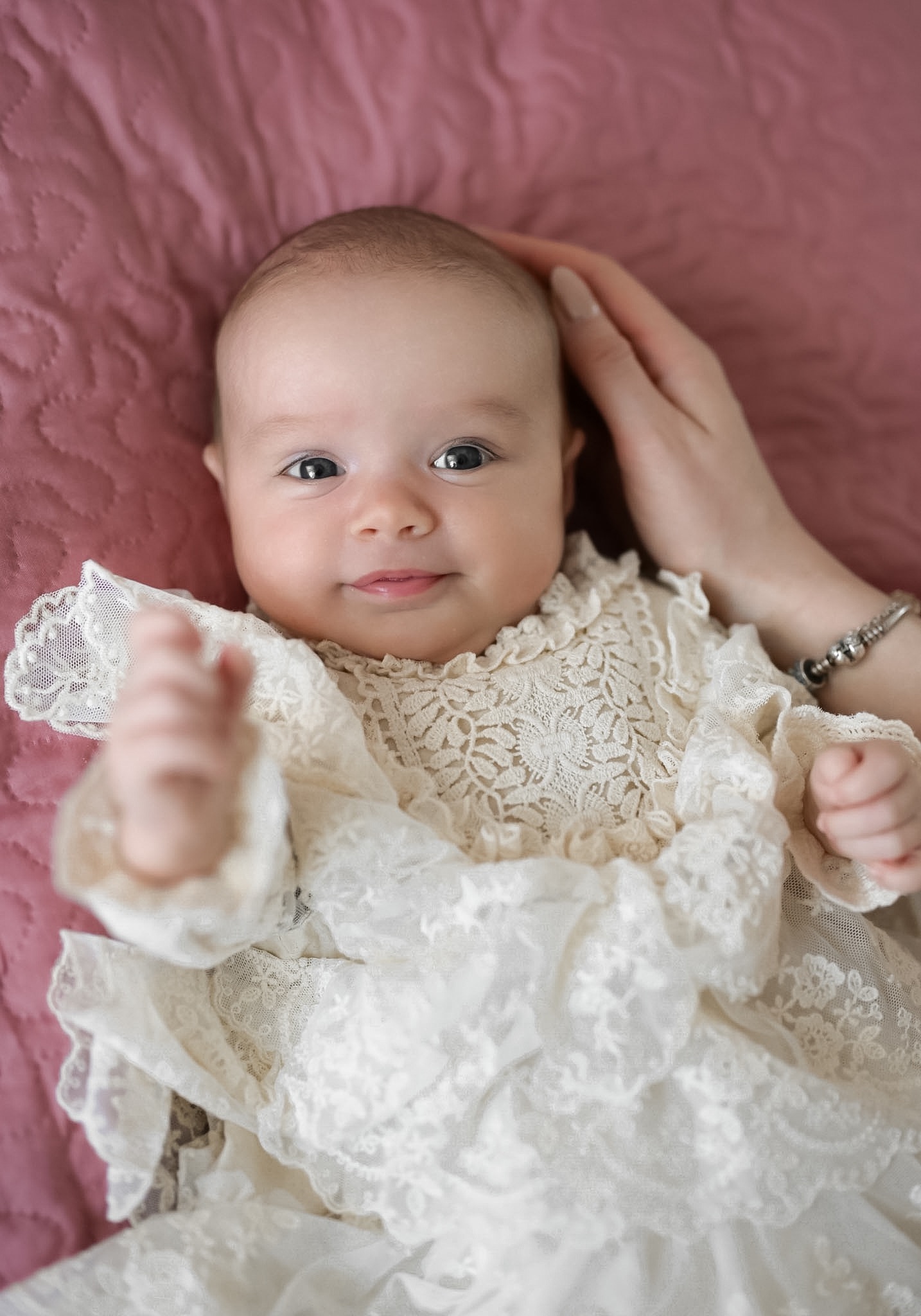 Urocze niemowlę w koronkowej sukience na różowym tle, delikatnie dotykane przez dłoń matki. Zdjęcie z Nagawczyny.