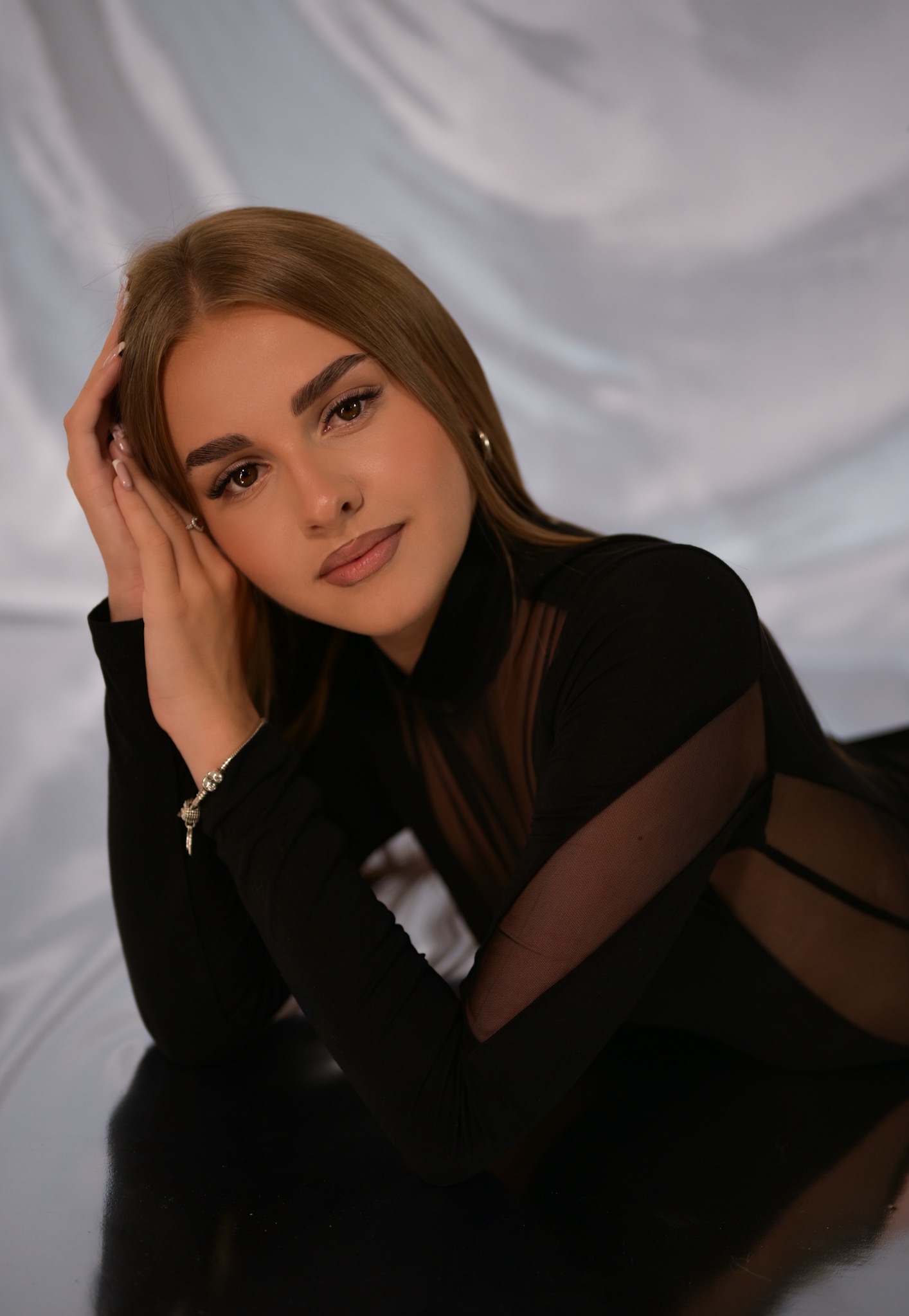 Portret kobiety w czarnym body z siateczkowymi wstawkami, leżącej na błyszczącej powierzchni z rękami pod głową, na tle białej, drapowanej tkaniny.